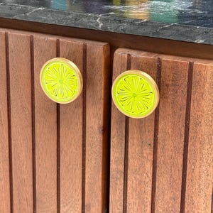 May include: Two circular cabinet knobs featuring a bright lime green citrus slice design with a gold-toned metal rim. The knobs are mounted on vertical slats of dark brown wood cabinetry, showcasing a detailed, textured fruit segment pattern.