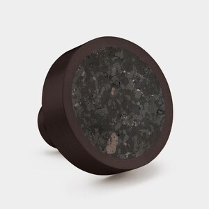 Puede incluir: Un pomo de mueble redondo con un marco de metal marr&oacute;n oscuro y una incrustaci&oacute;n circular de piedra moteada gris oscuro con motas minerales visibles. El pomo tiene un acabado mate liso en el borde exterior y una base cil&iacute;ndrica para montaje.