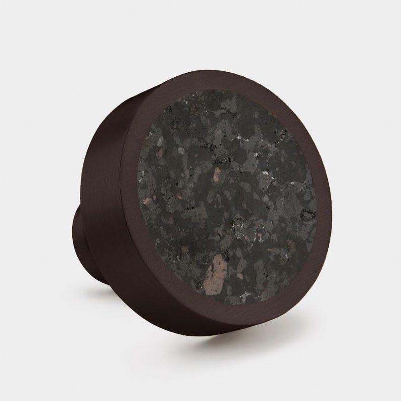 Puede incluir: Un pomo de mueble redondo con un marco de metal marr&oacute;n oscuro y una incrustaci&oacute;n circular de piedra moteada gris oscuro con motas minerales visibles. El pomo tiene un acabado mate liso en el borde exterior y una base cil&iacute;ndrica para montaje.