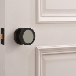 May include: A black door knob with a circular design and a textured edge is mounted on a white door. The knob has a flat, gray center. The door also features a black door latch.