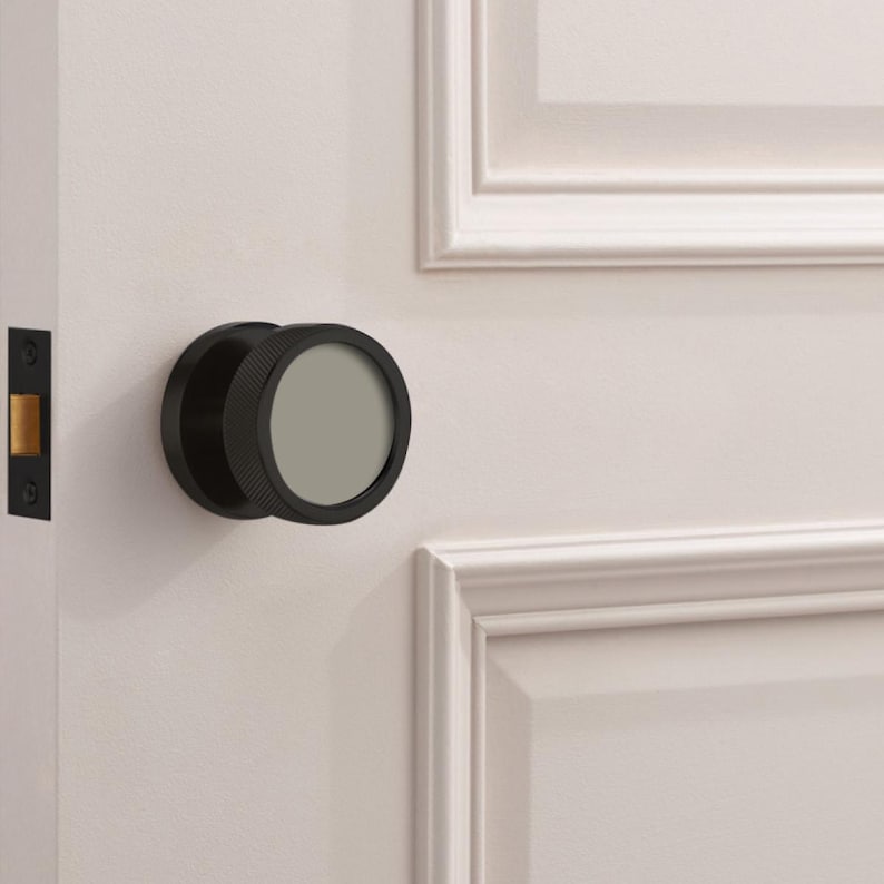 May include: A black door knob with a circular design and a textured edge is mounted on a white door. The knob has a flat, gray center. The door also features a black door latch.