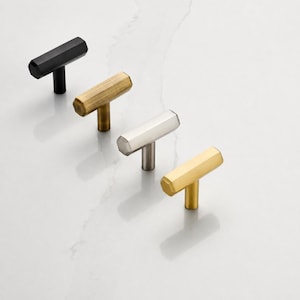 May include: Four T-shaped cabinet knobs arranged in a diagonal line. The knobs feature a hexagonal bar design and are finished in matte black, brushed antique brass, brushed nickel, and polished gold metal, each with a single cylindrical base.