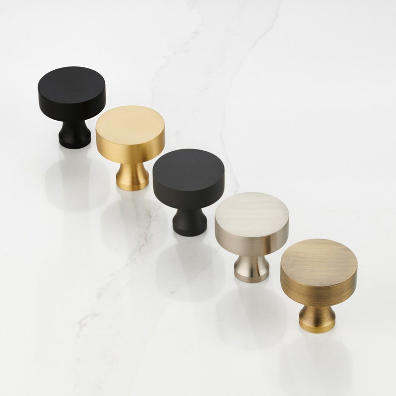 May include: Five round cabinet knobs in different finishes: black, gold, silver, and brass. The knobs have a simple, modern design.