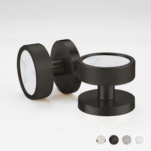 Puede incluir: Tres pomos de armario negros con diseño circular. Cada pomo presenta un centro blanco similar al mármol y un anillo exterior cilíndrico y estriado. Los pomos se muestran sobre un fondo neutro, mostrando su estética moderna.