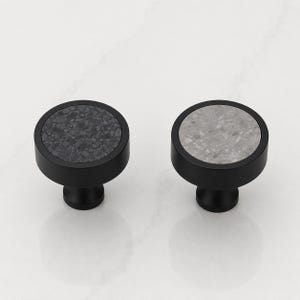 May include: Two round cabinet knobs with matte black metal bases. One knob features a dark charcoal gray speckled stone inlay, while the other knob features a light gray speckled stone inlay. Both knobs have a smooth, cylindrical profile.