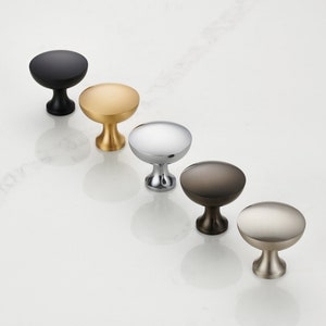 May include: An assortment of cabinet knobs in various finishes: black, gold, chrome, bronze, and brushed nickel. Each knob has a rounded top and a pedestal base. The knobs are arranged diagonally on a white surface, showcasing their different metallic appearances.