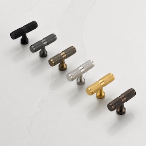 May include: A collection of six T-shaped cabinet knobs arranged in a diagonal line. Each knob features a cylindrical bar with a textured, cross-hatched knurled surface and a central mounting post. The finishes include matte black, dark bronze, antique brass, brushed silver, polished gold, and copper tones.