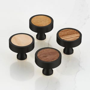 Puede incluir: Cuatro tiradores de armario negros con tapas circulares de madera en diferentes tonos de marrón claro y oscuro. Los tiradores tienen un acabado negro mate y una base redondeada de estilo pedestal. Los patrones de la veta de la madera son visibles.