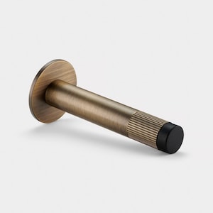 May include: A bronze-colored door stop with a circular base and a cylindrical body. The body has a ribbed texture on the end and a black rubber tip. The door stop is designed to prevent doors from slamming shut.