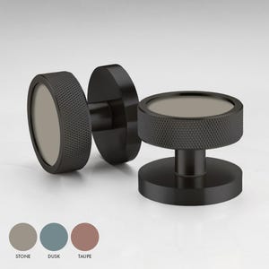 May include: Two black cabinet knobs with a textured finish. The knobs have a round, flat top with a circular inset in the center. The inset is available in three colors: stone, dusk, and taupe.