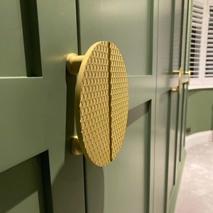 May include: A gold-colored cabinet door handle with a geometric pattern. The handle is round and has a split in the middle.