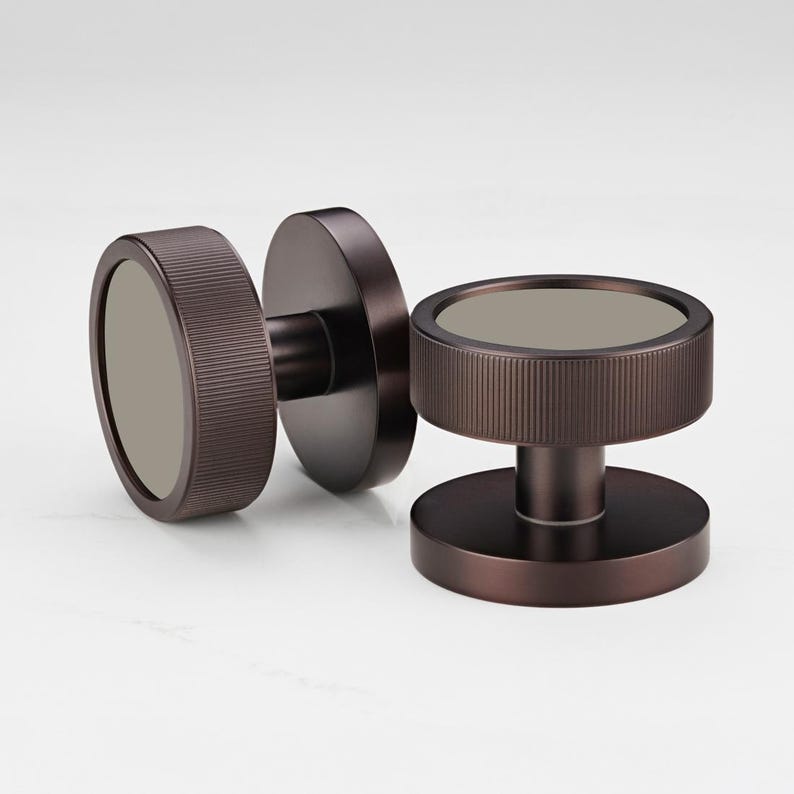 Puede incluir: Dos pomos de puerta de color bronce con un dise&ntilde;o circular. Cada pomo presenta un anillo exterior cil&iacute;ndrico estriado y un centro plano y circular. Los pomos se colocan sobre un fondo blanco, mostrando su est&eacute;tica moderna.