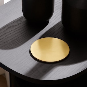 May include: A gold-colored, round coaster sits on a dark gray wooden surface. The coaster is made of a solid, reflective material. Two black, cylindrical objects are partially visible in the background, casting shadows on the surface.