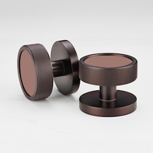 Puede incluir: Dos pomos de puerta de color bronce con un dise&ntilde;o circular. Los pomos tienen un anillo exterior cil&iacute;ndrico y estriado y un centro plano y circular. Los pomos est&aacute;n sobre un fondo blanco.