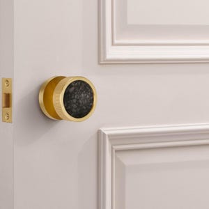 May include: A gold-toned door knob with a textured, dark gray center. The knob is round with a ridged edge and is mounted on a light-colored door. The door also features a gold-colored door latch.