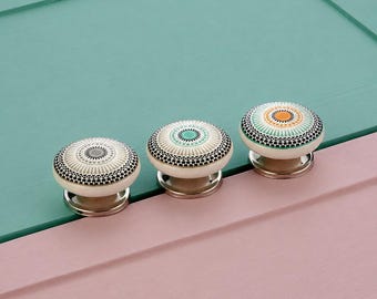 Può includere: Tre pomelli rotondi per armadietti in ceramica con superficie superiore piatta. Ogni pomello presenta un motivo dettagliato in stile mandala in nero, bianco e verde acqua, con il pomello a destra che include anche accenti arancioni. Base in metallo argentato.