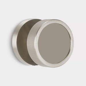 Może przedstawiać: Okrągłe lustro ścienne ze szczotkowanego metalu z żebrowaną krawędzią. Lustro ma płaską, szarą powierzchnię i jest zamontowane na białej ścianie. Projekt jest nowoczesny i minimalistyczny.