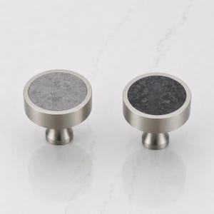 May include: Two circular cabinet knobs with a brushed silver metal finish. The left knob features a light gray speckled stone inlay, while the right knob features a dark charcoal black speckled stone inlay, both set within a smooth metal frame.