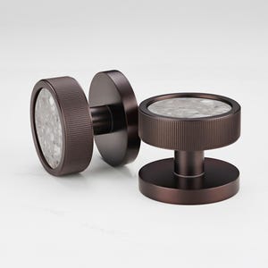 May include: Two bronze-colored cabinet knobs with a circular design. Each knob features a textured, ridged band around the edge and a speckled, gray and white stone center. The knobs are designed for easy installation and add a touch of elegance.
