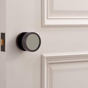 Puede incluir: Un pomo de puerta de bronce oscuro con un dise&ntilde;o circular y un centro gris plano. El pomo est&aacute; montado en una puerta blanca con un marco de puerta a juego y un pestillo de puerta de bronce oscuro.