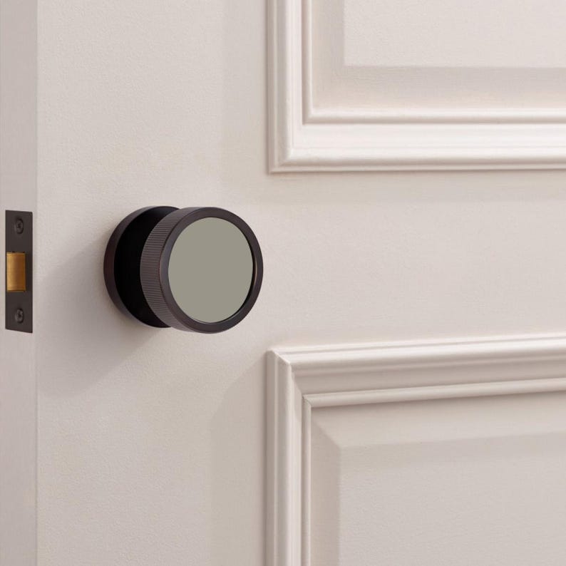 Puede incluir: Un pomo de puerta de bronce oscuro con un dise&ntilde;o circular y un centro gris plano. El pomo est&aacute; montado en una puerta blanca con un marco de puerta a juego y un pestillo de puerta de bronce oscuro.