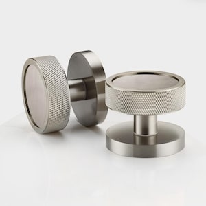 May include: Two brushed nickel door knobs. One knob is attached to a round plate, and the other is a standalone knob. The knobs have a textured, diamond-patterned grip around the edge and a smooth, flat top. The hardware is set against a white background.