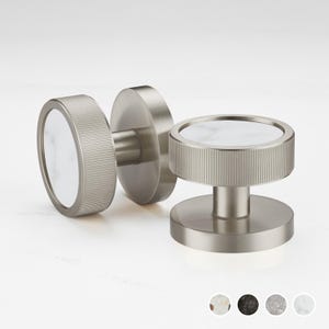 Puede incluir: Tres pomos de armario de níquel cepillado con incrustaciones de mármol blanco. Los pomos tienen un diseño circular con un borde estriado. Los pomos se muestran sobre una superficie blanca. Los pomos son de diseño moderno.