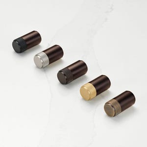 Puede incluir: Cinco tapones de botella cilíndricos con cuerpos marrón oscuro y varias tapas metálicas: negro, plateado, bronce oscuro, dorado y bronce. Los tapones están dispuestos en diagonal sobre una superficie de mármol blanco.