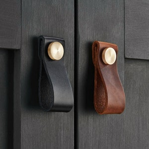 Puede incluir: Dos tiradores de armario de cuero con pomos de latón. Un tirador es de cuero negro y el otro de cuero marrón.