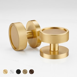 May include: Two gold-colored cabinet knobs with a circular design. The knobs have a ridged texture on the cylindrical portion and a smooth, flat surface on the circular face. The hardware is displayed against a white background.