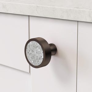 Puede incluir: Un pomo de armario redondo con un marco de metal bronce oscuro y una incrustaci&oacute;n circular con un patr&oacute;n de terrazo gris claro moteado. El pomo est&aacute; montado sobre una superficie blanca plana, mostrando un acabado liso y una base cil&iacute;ndrica.