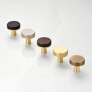 May include: Five cabinet knobs in various finishes: black, silver, brown, gold, and bronze. Each knob features a round, textured top and a brass-colored stem. The knobs are arranged diagonally on a white surface.