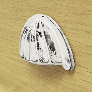 Könnte beinhalten: Weißer Keramik-Schubladengriff mit abgenutzter Oberfläche. Der Griff ist muschelförmig mit abgerundeter Oberseite und einem Loch für eine Schraube.