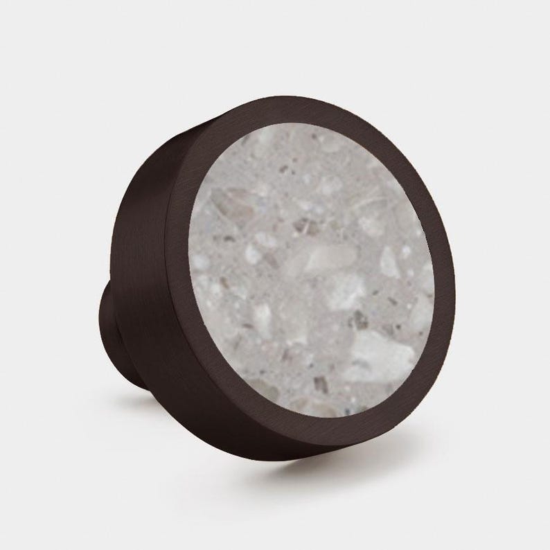 Puede incluir: Un pomo redondo para muebles con un marco de metal marr&oacute;n oscuro y una incrustaci&oacute;n circular de piedra terrazo gris claro. El pomo tiene un acabado mate suave en el borde exterior y una superficie texturizada y moteada en la cara frontal circular.