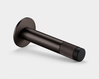 May include: A dark bronze cylindrical door stop featuring a circular wall-mounting plate and a textured, ribbed grip section near the end. The tip of the door stop is finished with a smooth, flat black rubber bumper for impact protection.