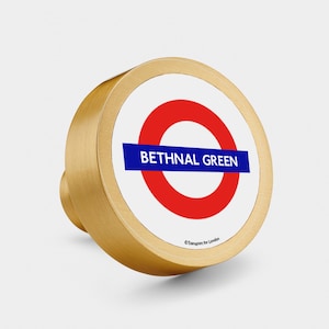 Puede incluir: Pomo redondo de color dorado con un círculo blanco y un anillo rojo, con el texto "BETHNAL GREEN" en letras azules y el símbolo de copyright con "Transport for London".