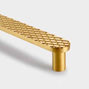 May include: A gold-toned cabinet pull with a diamond-shaped pattern. The pull has a rounded rectangular shape and a cylindrical base.