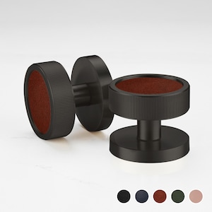 Pode incluir: Três botões de punho pretos com um design circular. Cada botão de punho apresenta um centro castanho escuro, semelhante a couro. Os botões de punho são exibidos numa superfície branca, com uma paleta de cores preta, azul marinho, castanha, verde e rosa na parte inferior.