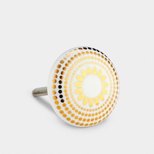 May include: White ceramic knob with a gold geometric design. The design features a circle of dots with a smaller circle of dots in the centre. The smaller circle has a flower-like design in the centre.