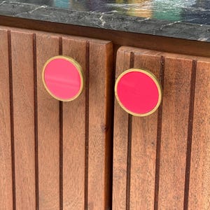 Op de afbeelding: Twee ronde kastknoppen met een felroze inleg in het midden, omringd door een dunne goudkleurige metalen rand. De knoppen zijn bevestigd op verticaal lattenhouten kastdeuren met een donkerbruine afwerking en een zichtbare houtnerf.