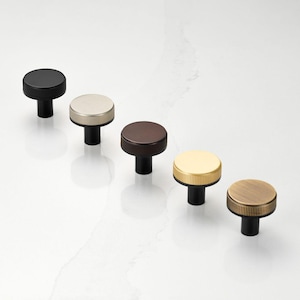 May include: A collection of five cabinet knobs in various metallic finishes. The knobs have a round, ridged top and a black stem. Colors include black, silver, bronze, gold, and brass, offering a range of design options.