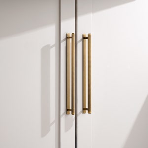 May include: Two gold cabinet pulls with a rectangular shape and rounded edges. The pulls are mounted on a white cabinet door.