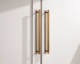 May include: Two long, vertical, brushed brass cabinet pulls with a rectangular profile. Each handle is mounted to a white door surface with two small, cylindrical supports, creating a clean, linear aesthetic with a metallic, golden-toned finish.
