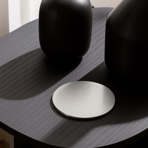 Puede incluir: Un posavasos redondo de metal cepillado sobre una mesa de madera oscura. Dos jarrones negros con acabado mate en el fondo. El posavasos es de diseño sencillo y moderno, ideal para proteger superficies.