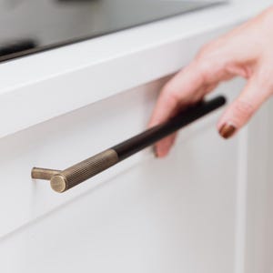 Op de afbeelding: Een witte kast met een zwarte en messingkleurige handgreep. De handgreep is een lange, horizontale staaf met een geribbelde textuur aan het uiteinde en een gladde, donkere afwerking.