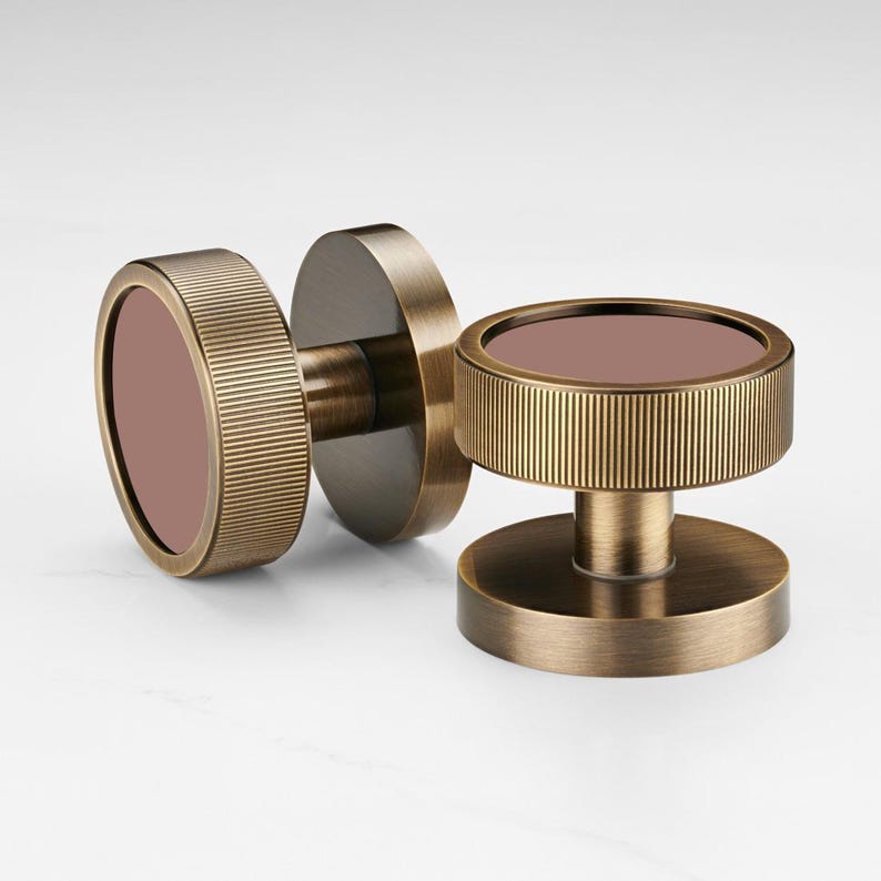 K&ouml;nnte beinhalten: Zwei antike Messing-Schrankkn&ouml;pfe mit kreisf&ouml;rmigem Design. Jeder Knopf hat einen geriffelten, zylindrischen Au&szlig;enring und eine flache, kreisf&ouml;rmige Mitte in einem ged&auml;mpften Rosaton. Die Kn&ouml;pfe sind vor einem wei&szlig;en Hintergrund platziert.