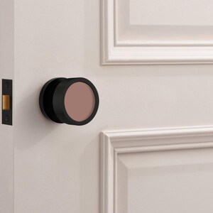 May include: A black door knob with a circular, muted rose-colored center. The knob is mounted on a white door with a matching white door frame. The door also features a black door latch.