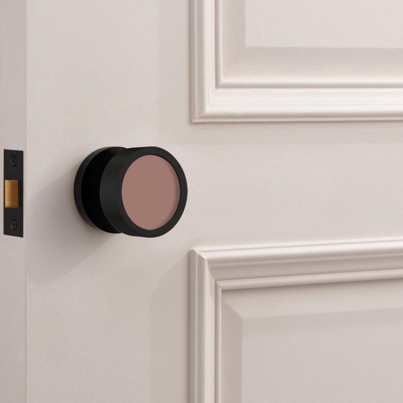May include: A black door knob with a circular, muted rose-colored center. The knob is mounted on a white door with a matching white door frame. The door also features a black door latch.