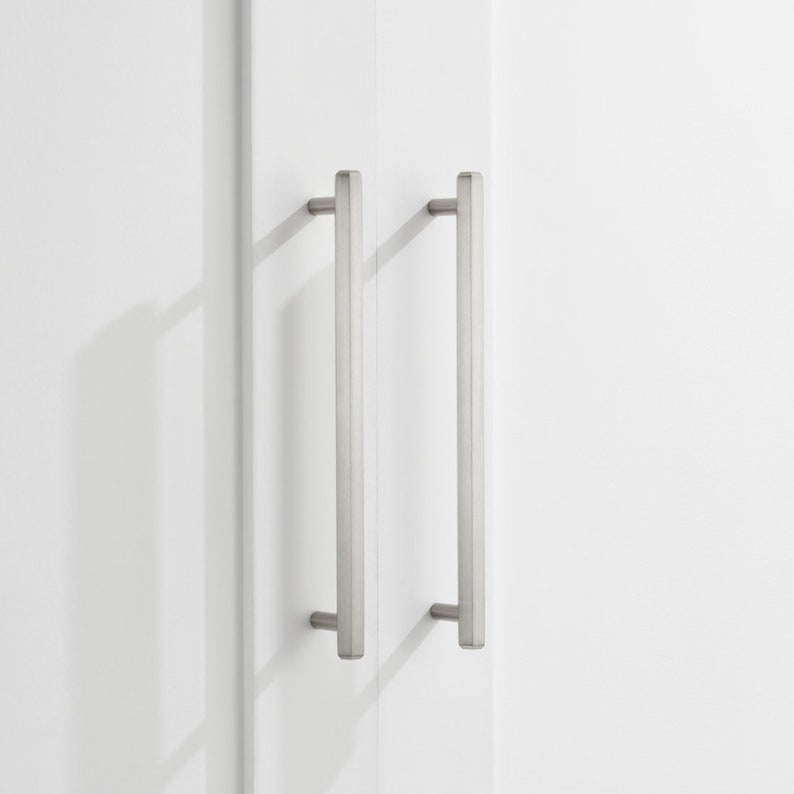 May include: Two brushed nickel cabinet pulls with a rectangular shape. The pulls are mounted on a white cabinet door.