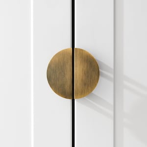 Op de afbeelding: Close-up van een witte kastdeur met een grote, ronde handgreep in antiek messing. De handgreep is in tweeën gesplitst en op de deur gemonteerd, met een zwarte verticale lijn in het midden.